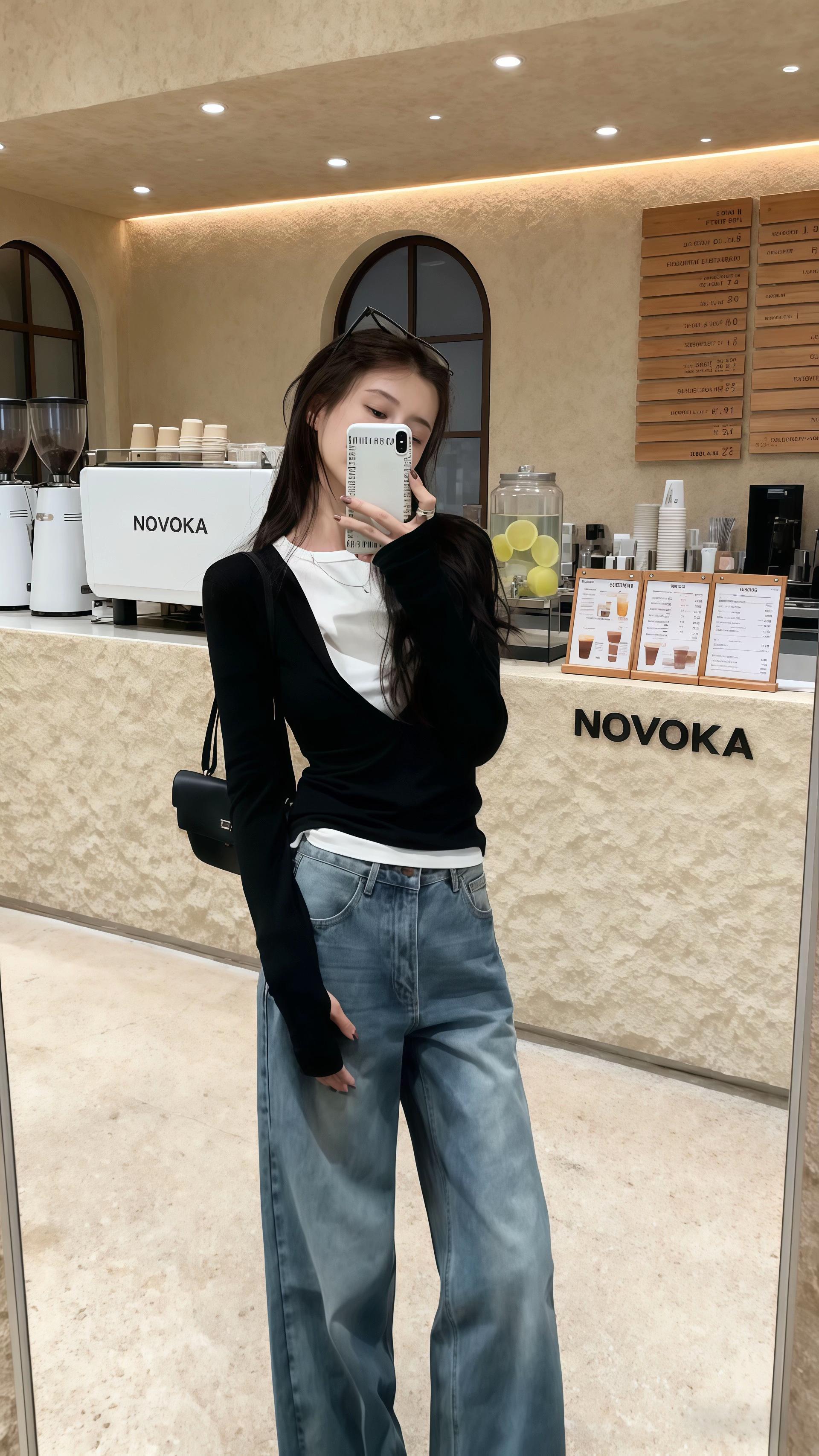 女生在一家名为“NOVOKA”的咖啡馆内自拍。她留着黑色长发,头顶架着一副墨镜,妆容精致,正拿着白色手机看向镜头。穿搭上,她内搭一件白色基础款T恤,外搭黑色交叉V领长袖上衣,勾勒出优雅的颈部与肩部线条;下身搭配一条浅蓝色的阔腿牛仔裤,宽松的版型尽显休闲随性;斜挎着一个黑色的小方包,简约又时尚。 背景的咖啡馆环境温馨,暖黄色的灯光、带有拱形设计的窗户,以及摆放着咖啡器具、柠檬片和菜单的吧台,营造出舒适惬意的氛围。整体穿搭简约又不失设计感,适合日常出街或休闲聚会等场合。#穿搭ootd